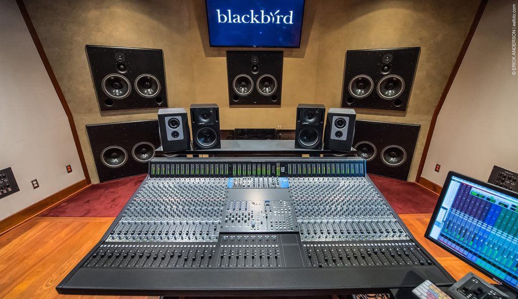 Dolby Atmos mixing room with large console, level meter display, multiple monitor speakers including three soffit-mounted front monitors, surround speakers, display showing Blackbird logo, red carpeted floor.