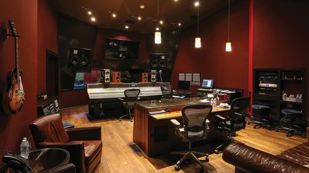 Control room with mixing console, studio monitors in soffit, guitars on wall, leather seating, pendant lights, red walls, production desk area.