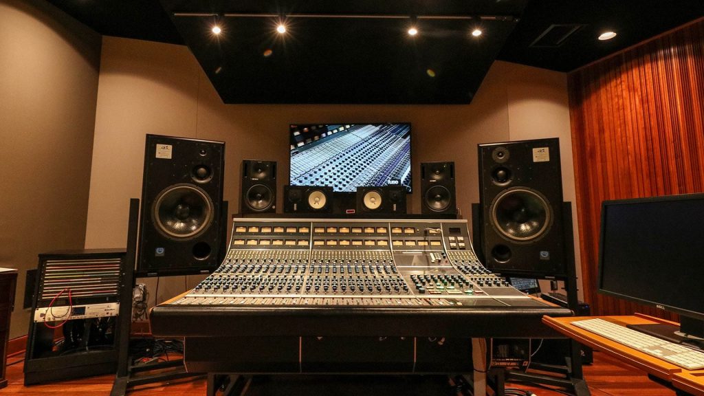 Control room with large console, monitor array including Genelec and Yamaha NS-10Ms, large display showing console image, orange wood acoustic treatment visible right.