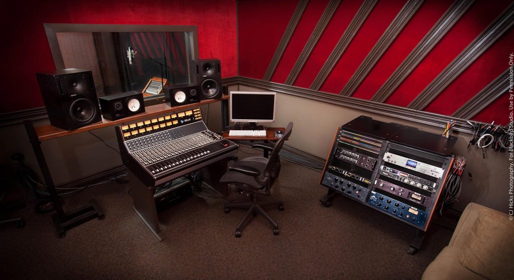 Control room with API console and meter bridge, office chair, computer monitor, window to vocal booth showing red curtains, multiple studio monitors including Yamaha NS-10Ms, red and striped acoustic treatment on ceiling, outboard gear rack right.