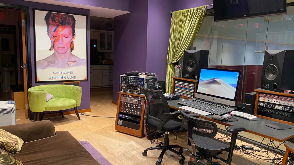 Control room with David Bowie Aladdin Sane poster, yellow-green velvet chair, production desk with iMac, MIDI controller, studio monitors, outboard gear racks, view to kitchen, purple walls.