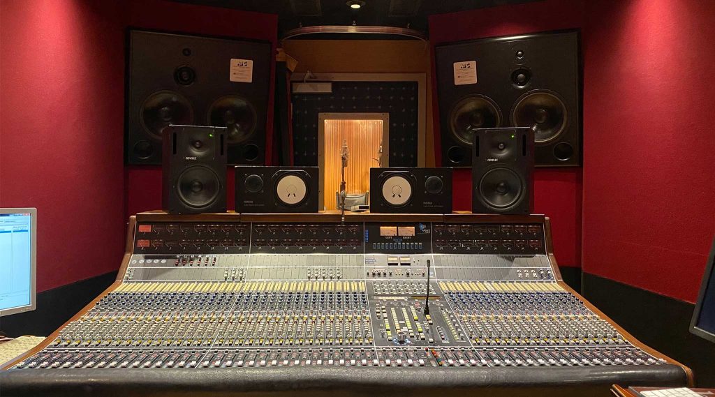 Mixing console with Genelec and Yamaha NS-10M monitors, VU meter bridge, window into wood-paneled vocal booth with microphone visible.